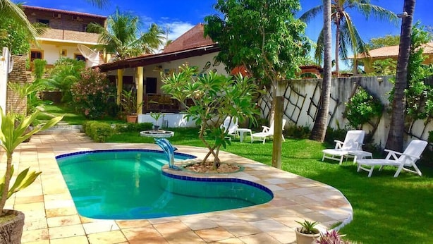 Une piscine à bord incurvé, entourée d’une terrasse carrelée et d’une pelouse agrémentée de chaises longues blanches.