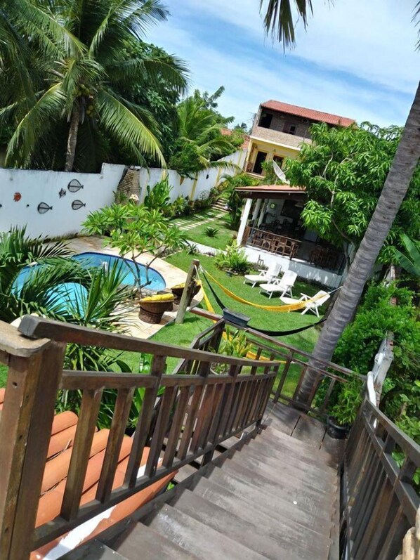 A wooden staircase leading to a pool area with a hammock, lounge chairs, and a white building with a red roof. - Pousada São Pedro Flecheiras (Trairi)
