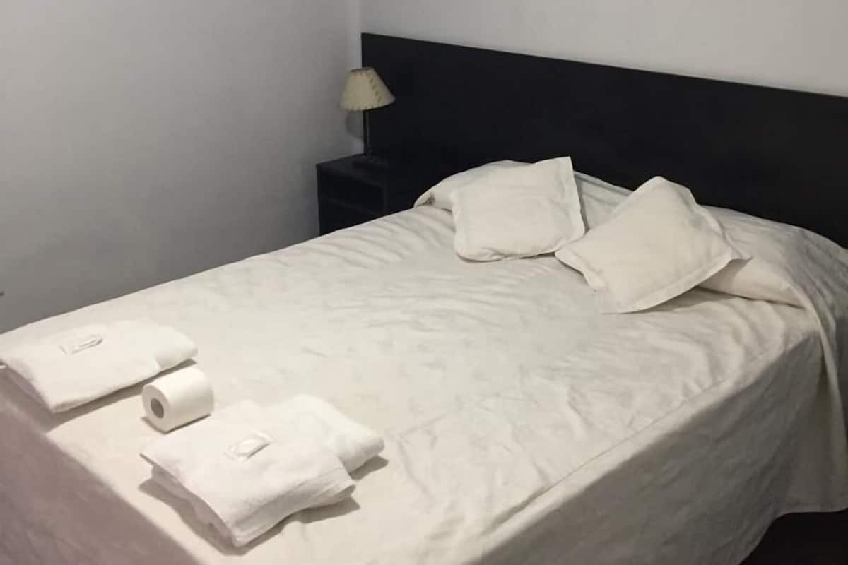 A neatly made bed with white linens and pillows, a small bedside table with a lamp, and a framed picture on the wall.