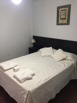 Una cama bien hecha con sábanas y almohadas blancas, una mesita de noche con lámpara y un cuadro en la pared.