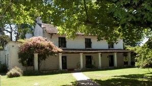 Een wit huis met een pannendak, omringd door een verzorgd gazon en een grote boom die schaduw biedt.
