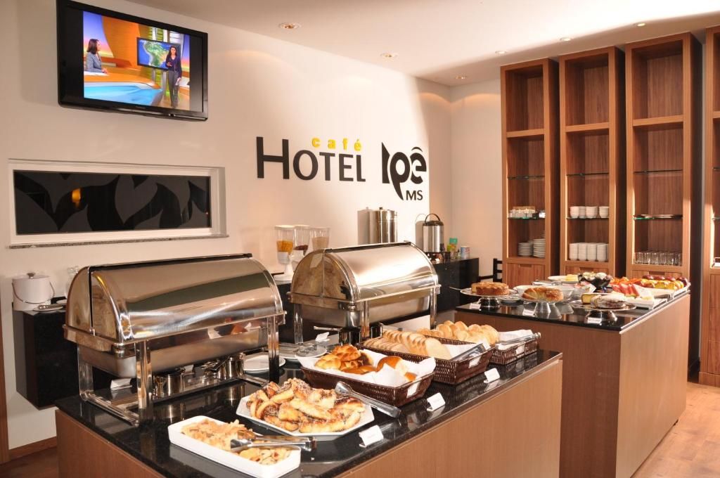 A hotel breakfast buffet with various food items, a television displaying a news program, and a wall sign reading 'café HOTEL L'OPE MS'.
