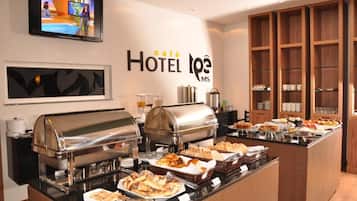 A hotel breakfast buffet with various food items, a television displaying a news program, and a wall sign reading 'café HOTEL L'OPE MS'.