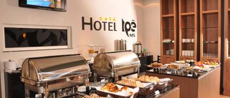 Un bufé de desayuno de hotel con diversos alimentos, un televisor mostrando un noticiero y un letrero en la pared que dice 'café HOTEL L'OPE MS'.