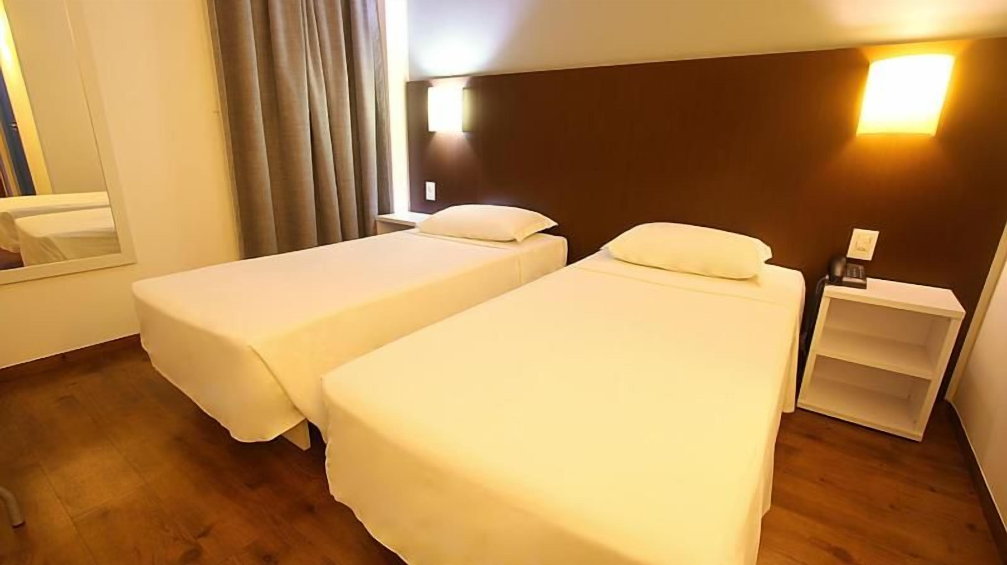 A hotel room with two single beds, a bedside table, a nightstand, a mirror, and a window with curtains.