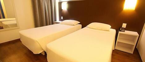 A hotel room with two single beds, a bedside table, a nightstand, a mirror, and a window with curtains.