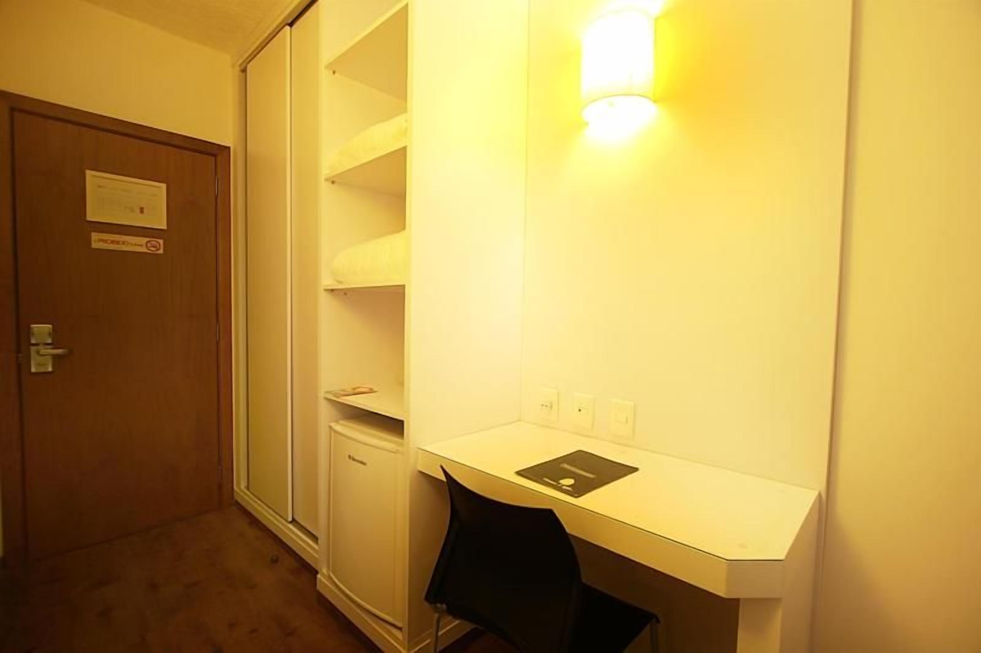 A hotel room with a wardrobe, a desk with a chair, and a wall-mounted light fixture.