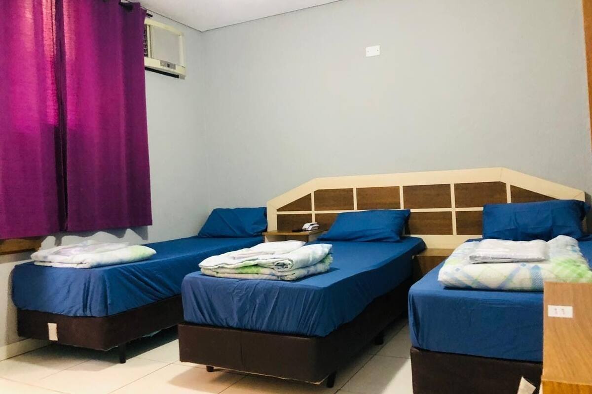 Une chambre avec trois lits, chacun doté de draps et d’oreillers bleus, et un rideau violet sur le côté gauche.