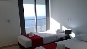 Ein Hotelzimmer mit zwei Betten, einem großen Fenster mit Meerblick, einem Nachttisch mit Lampe und roten Akzenten an den Betten.