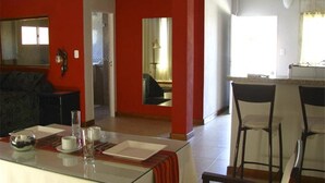 Un comedor con una mesa de vidrio, sillas y una mesa decorada con un mantel rojo. Se ve una cocina con mostrador y sillas al fondo.