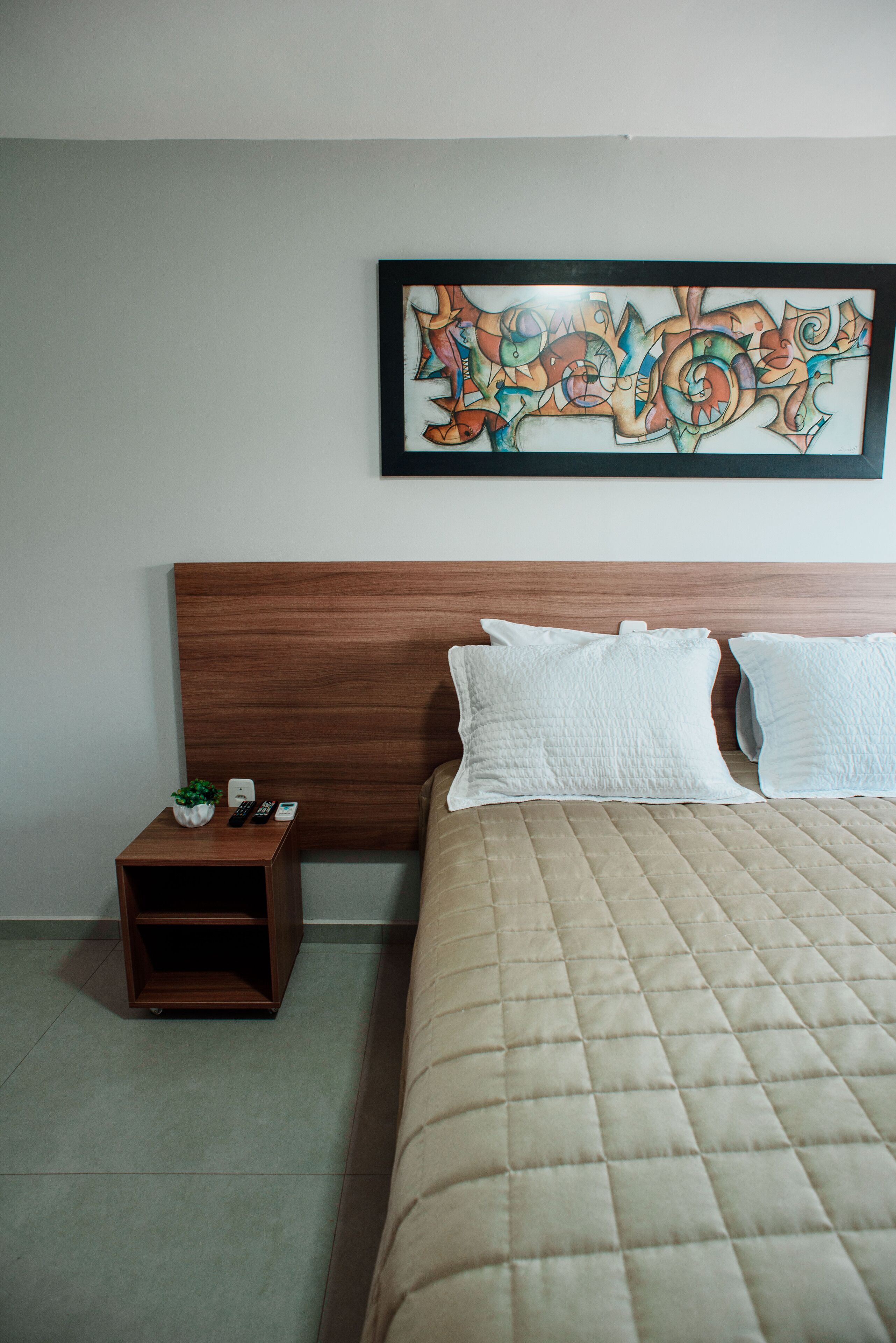 A hotel room with a wooden headboard, a quilted bedspread, a bedside table with a plant, and a framed abstract artwork on the wall.