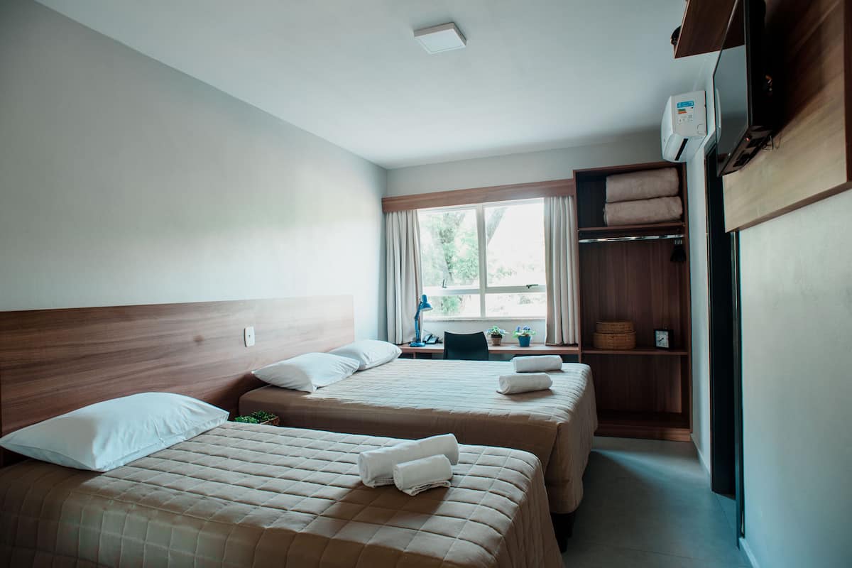 A hotel room with two beds, a desk with a chair, a window with curtains, and a wardrobe.