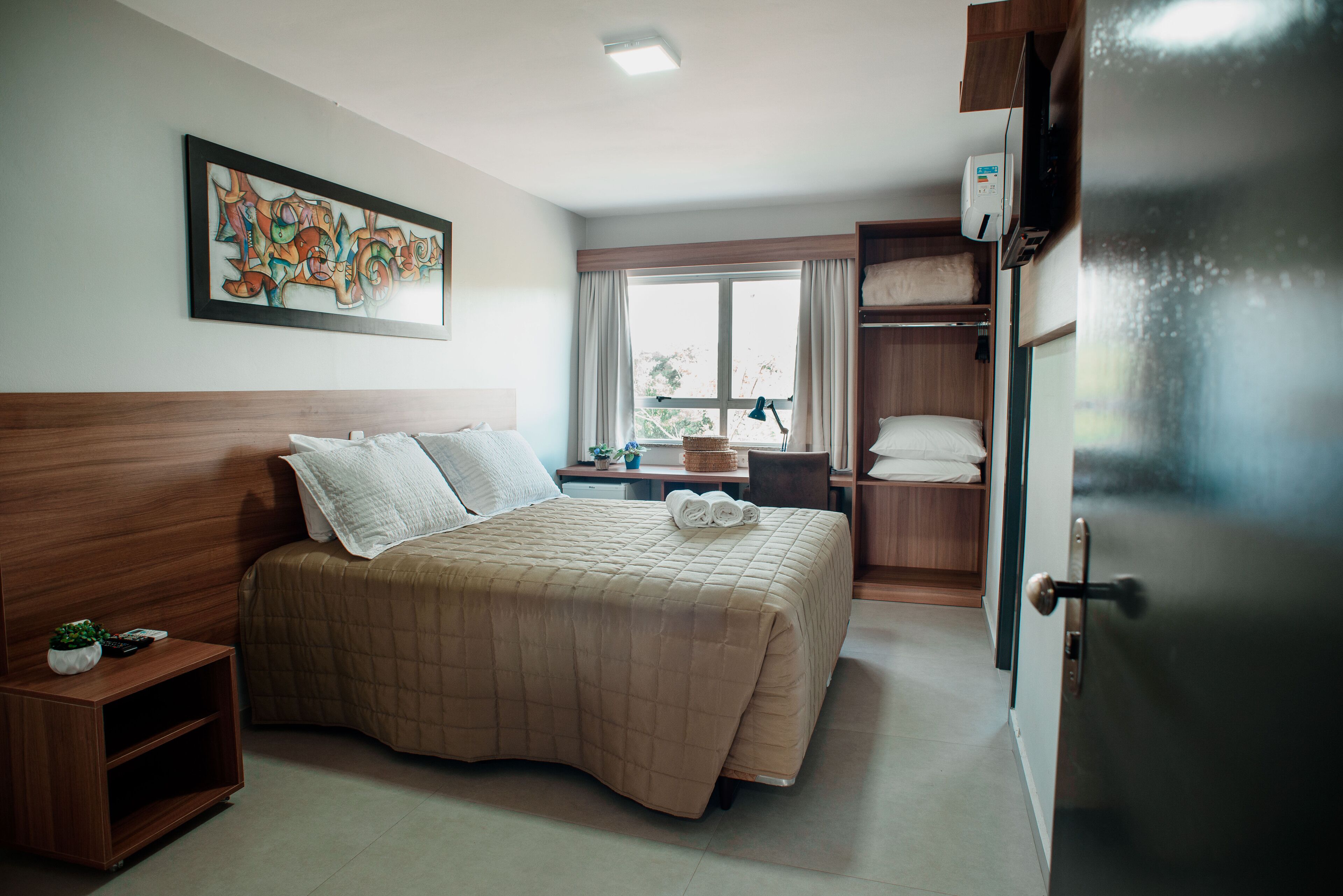 Una habitación de hotel con cama, mesita de noche, ventana, un cuadro y una puerta.