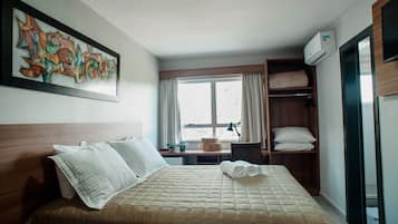 Et hotelværelse med seng, vindue, skrivebord, stol, hylder med puder og en vægmonteret aircondition.