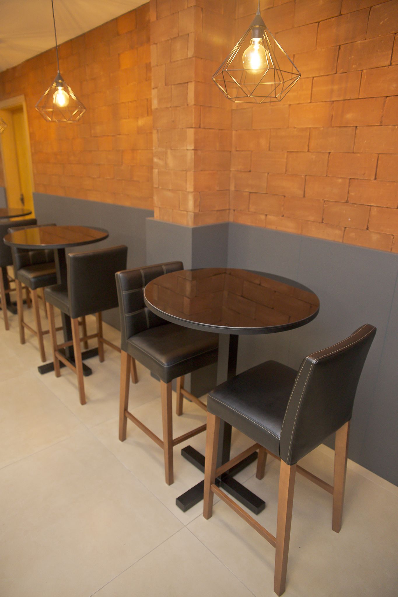 Un interior de restaurante con mesas redondas, sillas de cuero negro, paredes de ladrillo y lámparas colgantes.