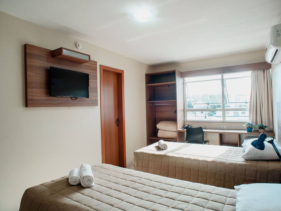 Habitación de hotel con dos camas, un televisor montado en la pared, un escritorio con una silla y una ventana con cortinas.