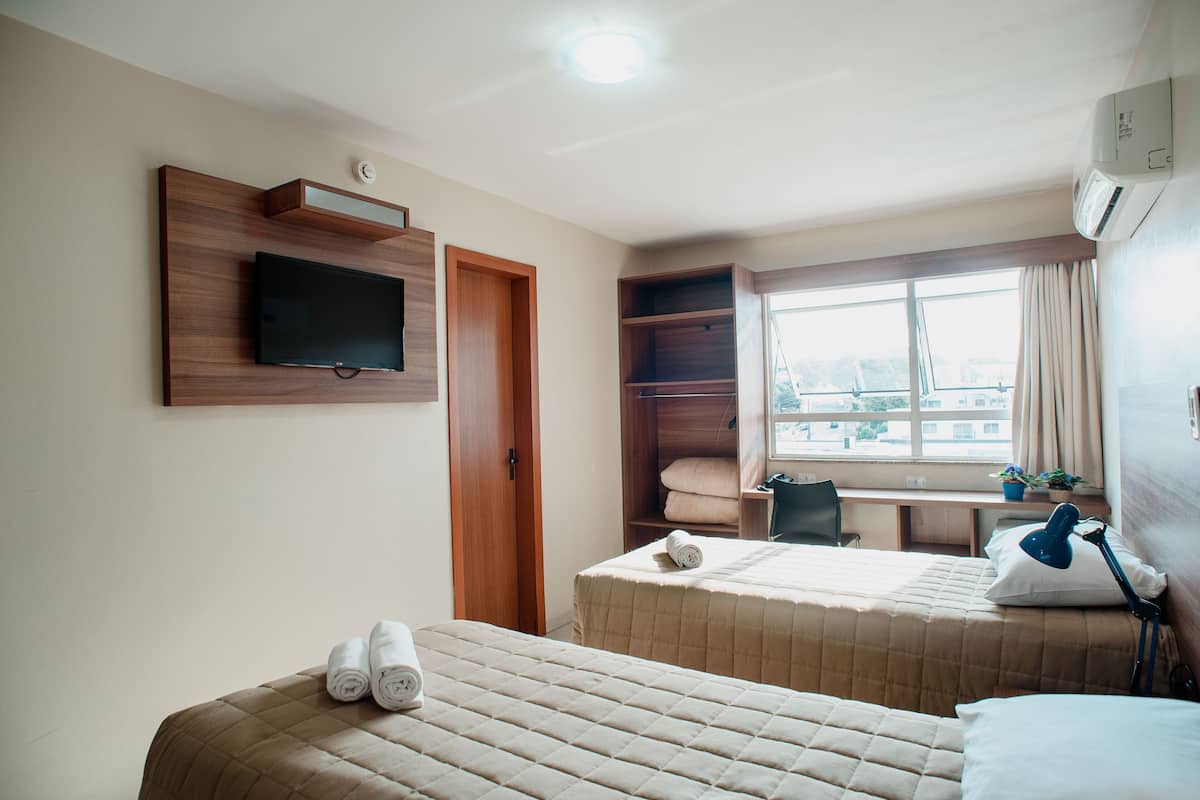 A hotel room with two beds, a TV mounted on the wall, a desk with a chair, and a window with curtains.