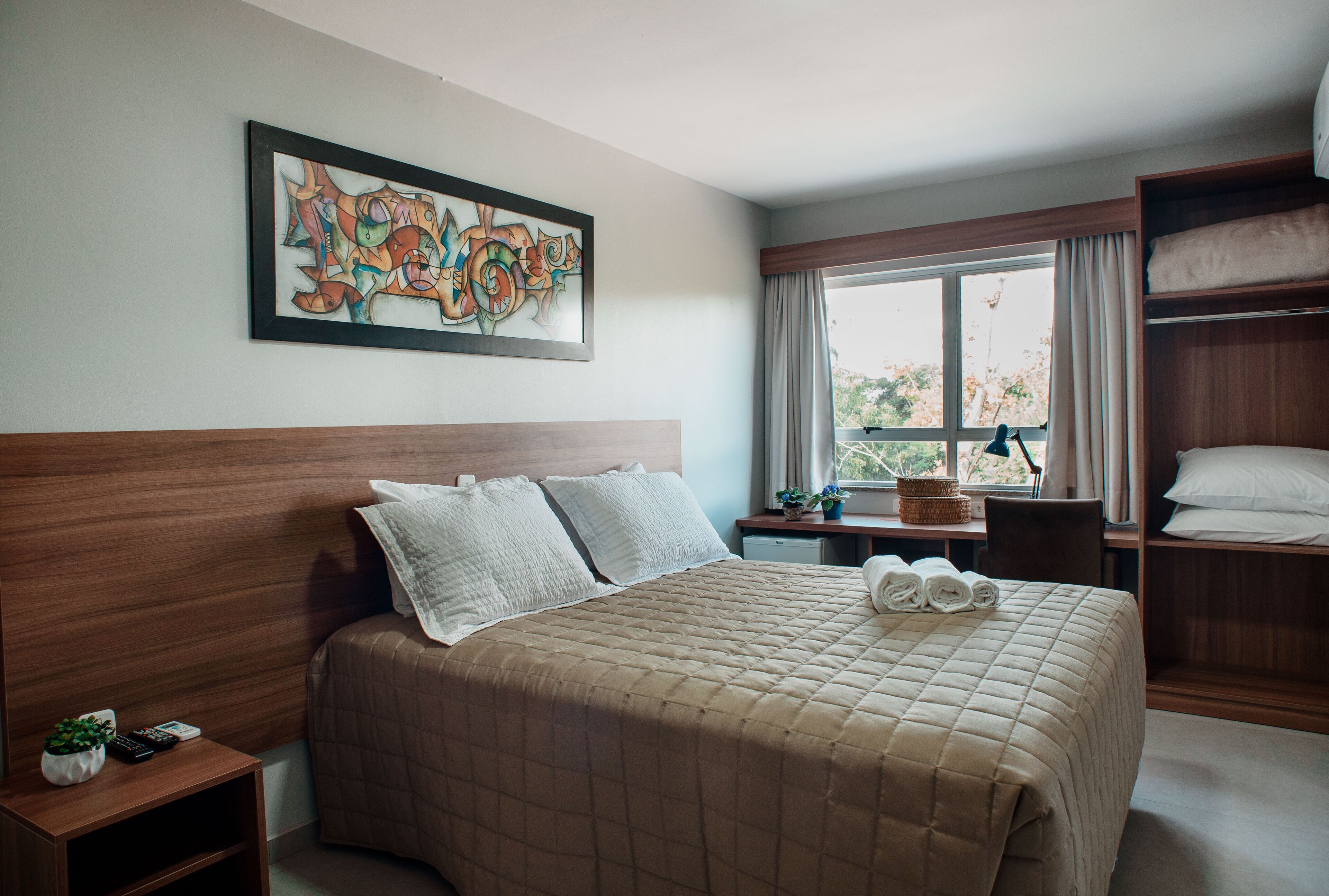 Una habitación de hotel con una cama grande, un cabecero de madera, un cuadro en la pared, una ventana con cortinas y un armario de madera.
