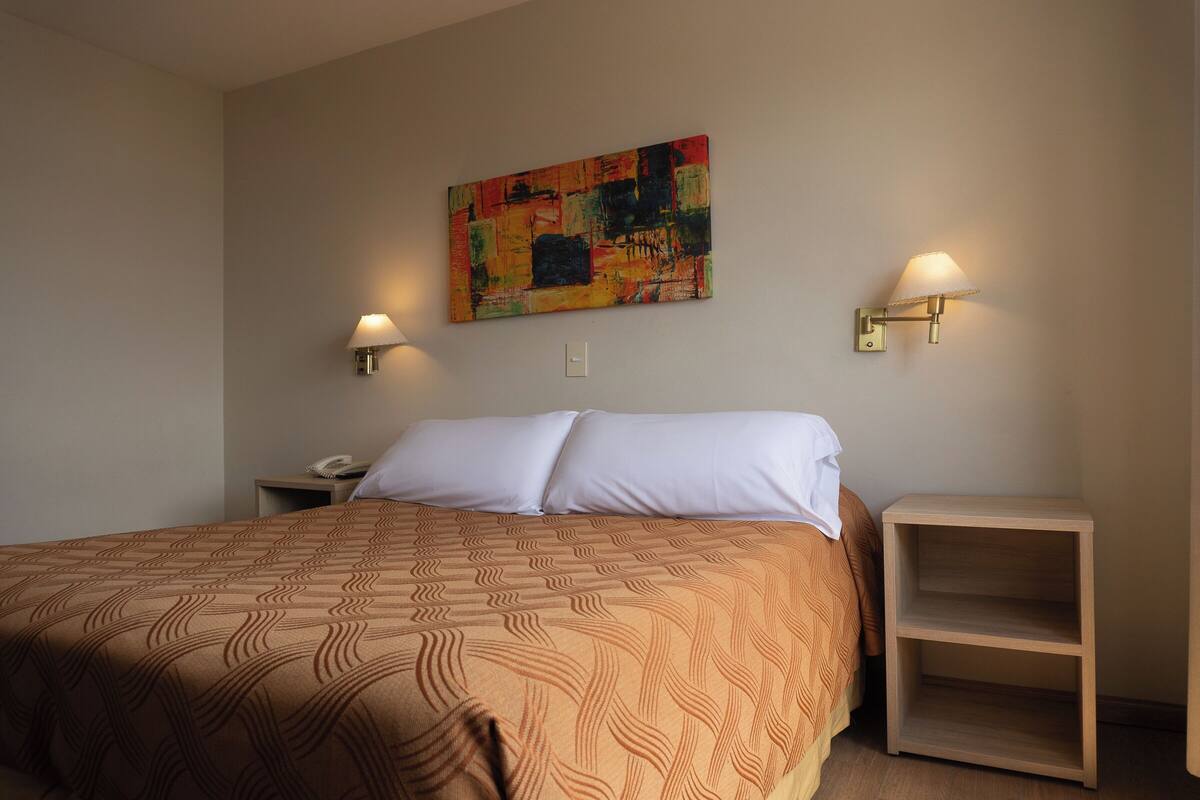 Ein Hotelzimmer mit einem großen Bett, zwei Nachttischlampen, einem Nachttisch und einem farbenfrohen abstrakten Gemälde an der Wand.