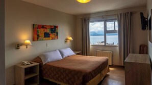 Ein Hotelzimmer mit Bett, Nachttischlampen, einem Bild an der Wand, einem Fenster mit Meerblick und einem Schreibtisch mit Stuhl.
