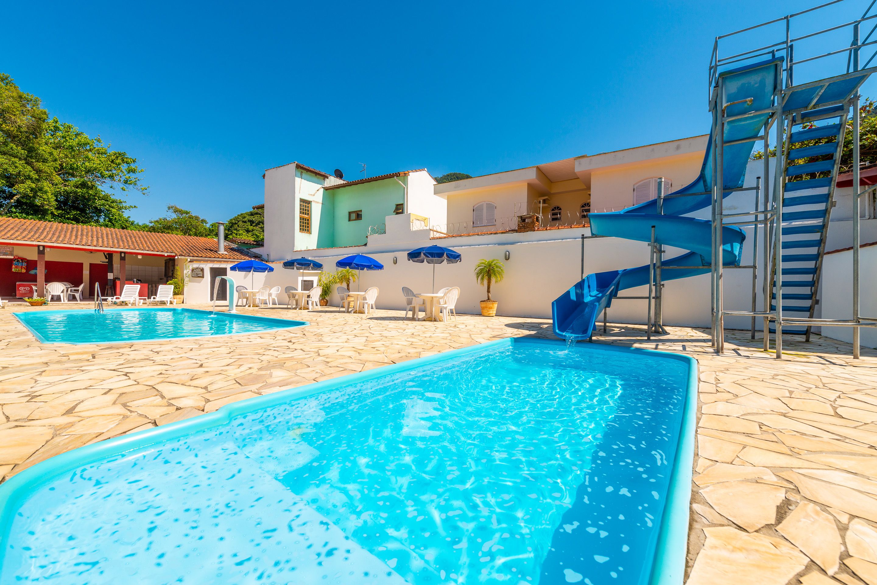 Una piscina amb tobogan, paviment de pedra i zona d'estar exterior amb ombrelles.