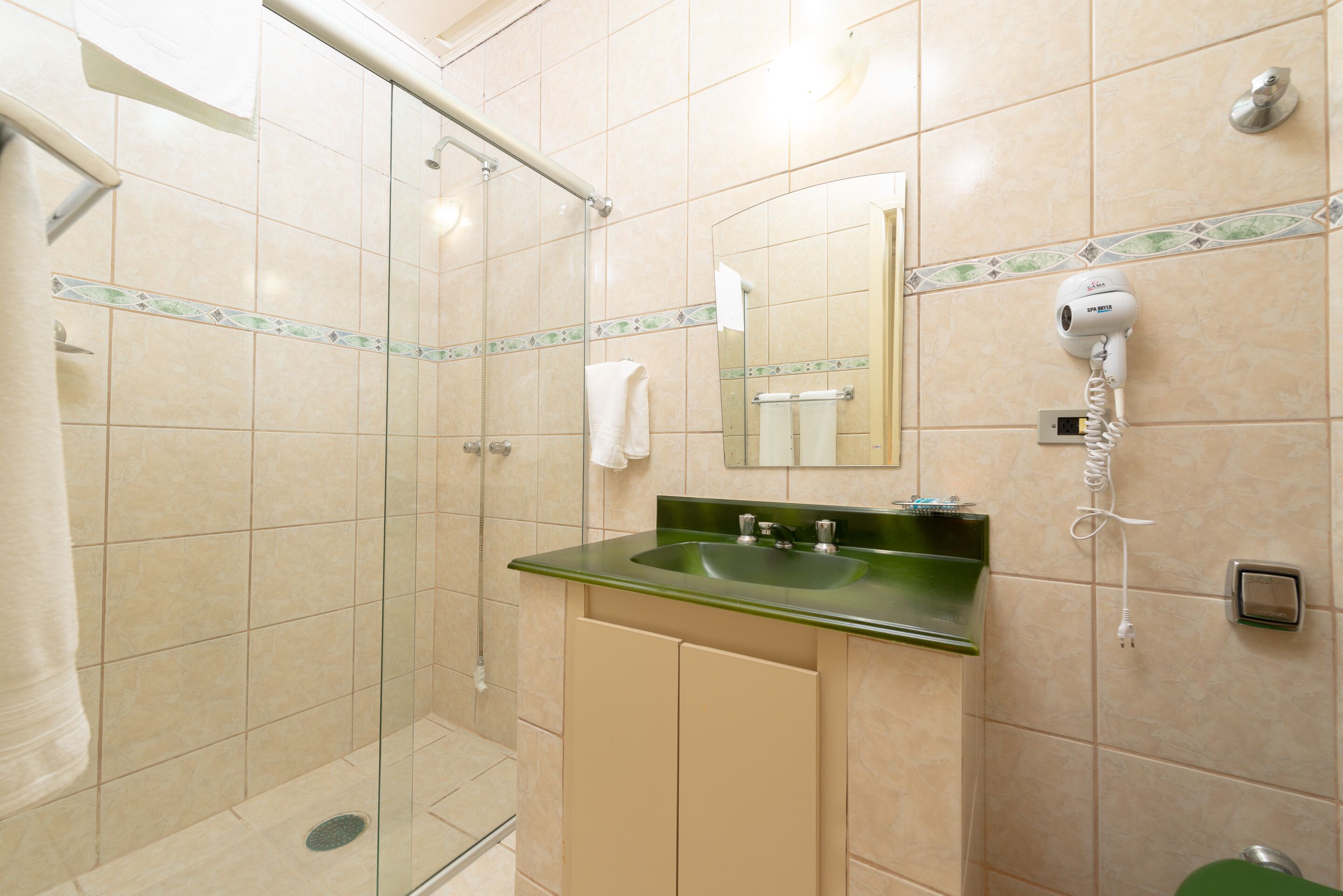Un baño con ducha de vidrio, un lavabo verde y un secador de pelo montado en la pared.