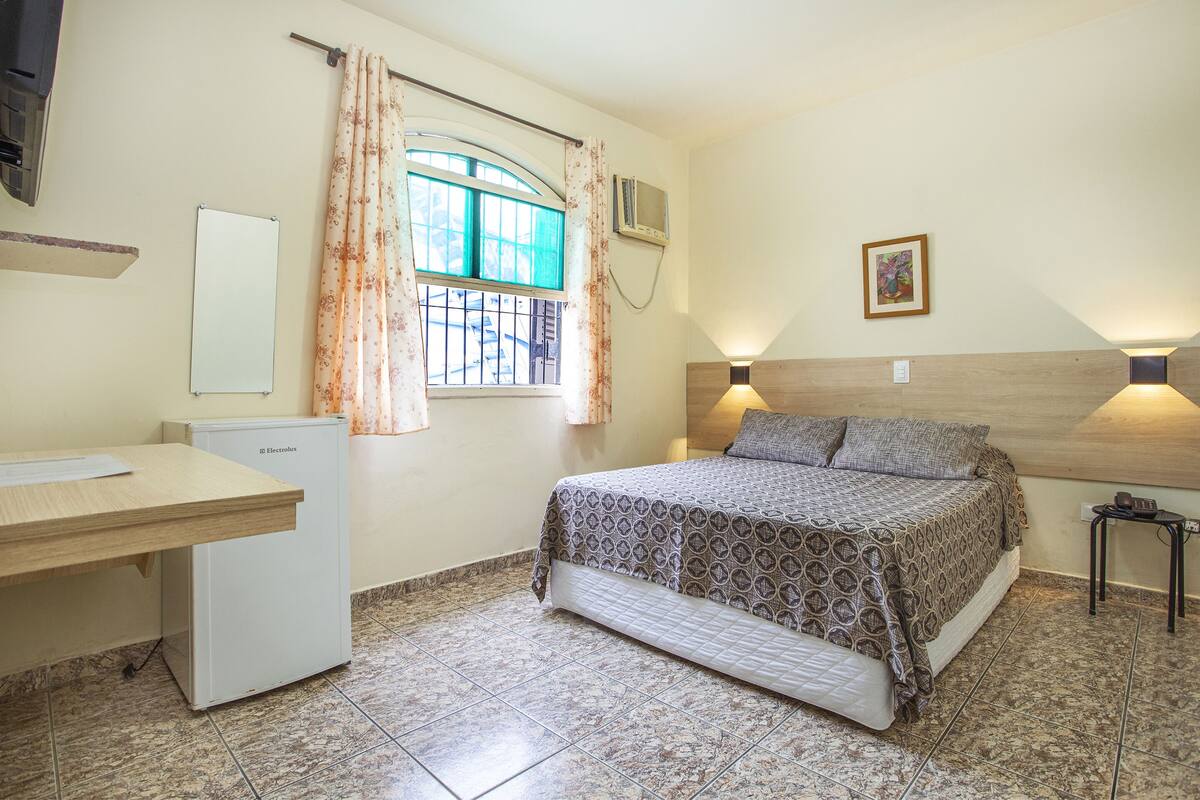 A bedroom with a bed, a desk, a refrigerator, a window with curtains, a wall-mounted air conditioner, a bedside lamp, and a framed picture on the wall.