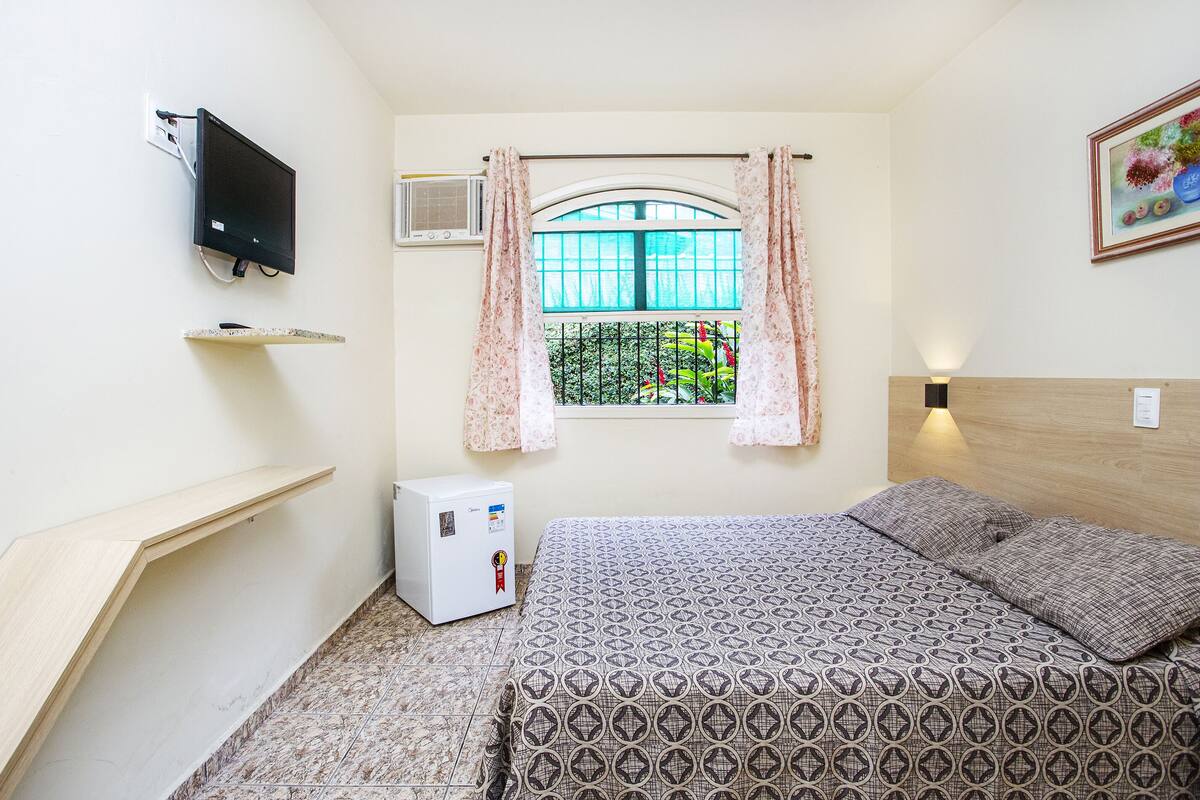 A small, neatly arranged bedroom with a bed, a TV mounted on the wall, a small air conditioner, and a window with curtains.