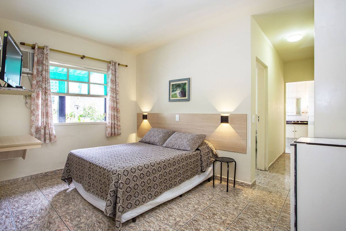 Un dormitorio con cama, una mesita, un televisor y una ventana con cortinas.