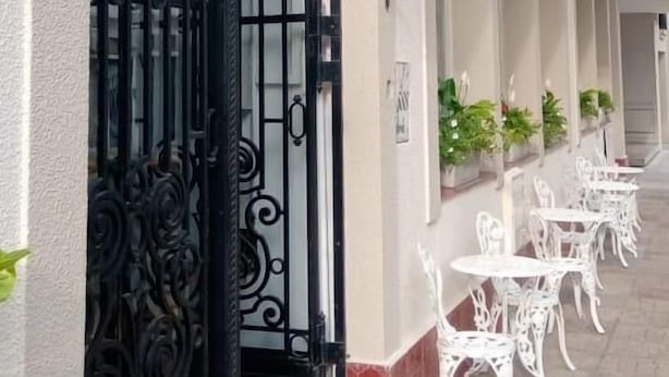 Entrada de un hotel con una reja de hierro forjado negro, mobiliario blanco para exteriores y un cartel.