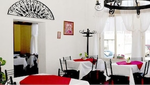 Comedor con mesas cubiertas de manteles blancos y caminos de mesa rojos, sillas negras y un ventilador de techo decorativo.