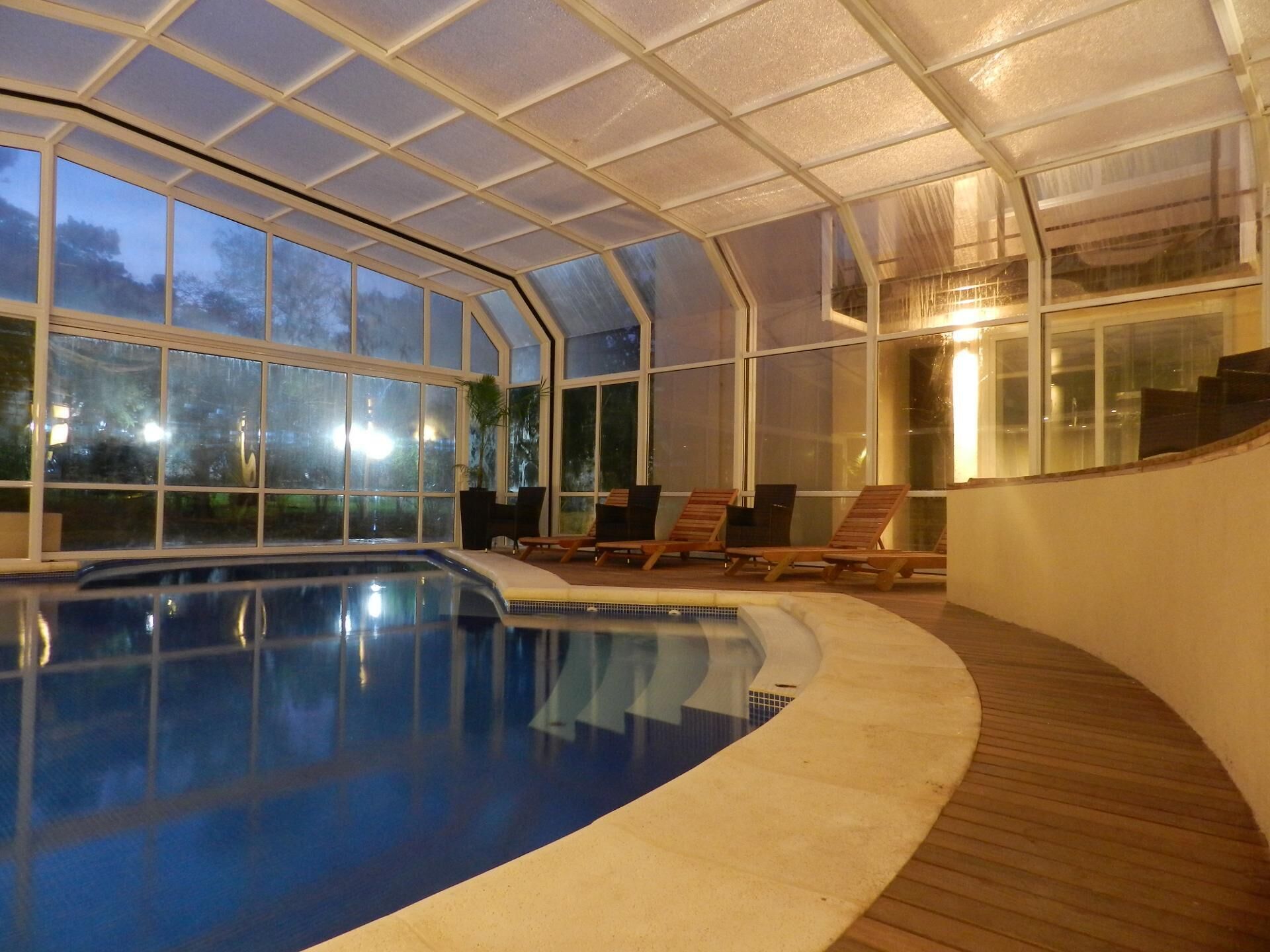 An indoor swimming pool with clear glass walls, wooden deck, and lounge chairs.