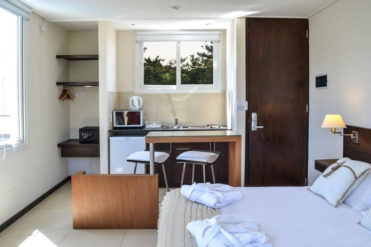 A compact hotel room with a kitchenette, a bed with white linens, and a small seating area.