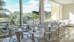 Un comedor moderno con sillas y mesas blancas, ventanas amplias que ofrecen vistas a la vegetación y un cielo despejado.