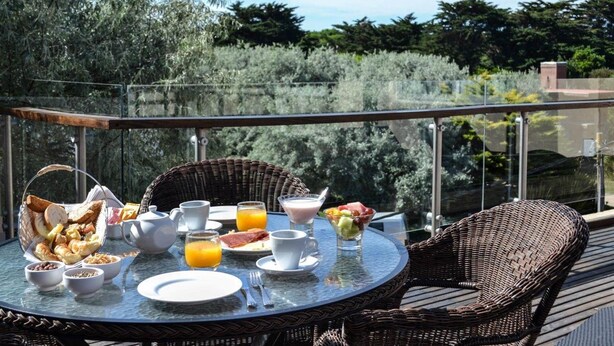 Una mesa redonda con alimentos para el desayuno, incluyendo pan, fruta y bebidas, en un balcón con sillas de mimbre y barandilla de vidrio.
