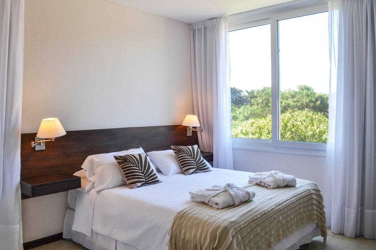 A hotel room with a large bed, two bedside lamps, a window with curtains, and a view of greenery outside.