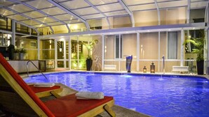 Una amplia piscina cubierta con techo de cristal, sillones reclinables rojos y una pequeña cascada.