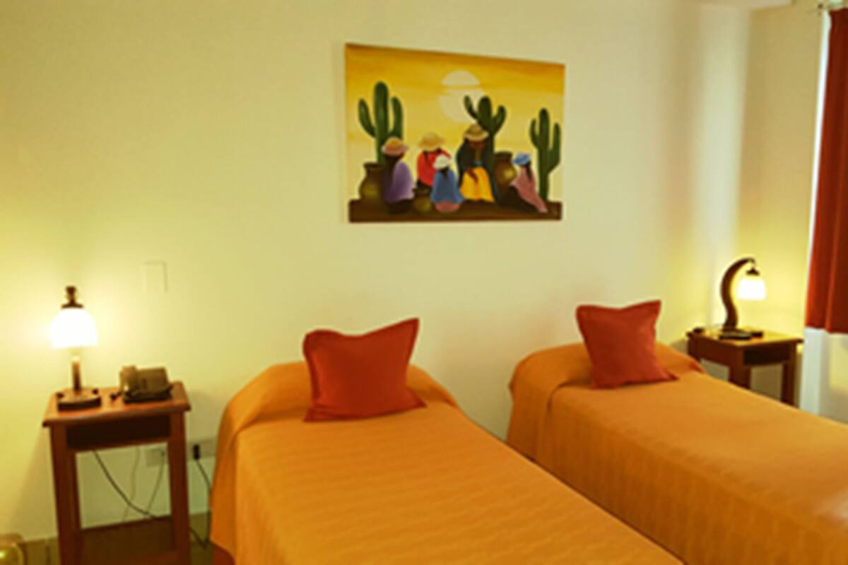 A hotel room with two beds, a painting of cacti, a lamp, and a window with curtains.
