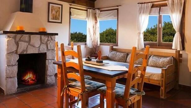 A cozy room with a stone fireplace, wooden furniture, and large windows with curtains. - Cabañas Puerto Delfín (Mar de las Pampas)