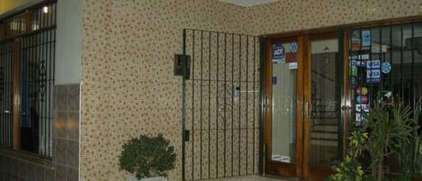 A building entrance with a tiled wall, a metal gate, a small potted plant, and a door with various stickers.