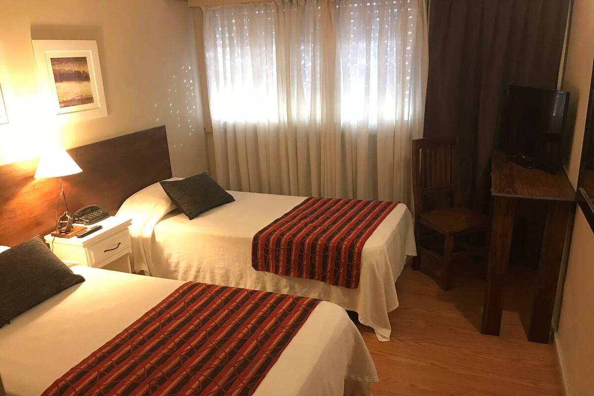 Quarto de hotel com duas camas, uma mesa de cabeceira com luminária e telefone, uma TV sobre um suporte de madeira e uma janela com cortinas leves.