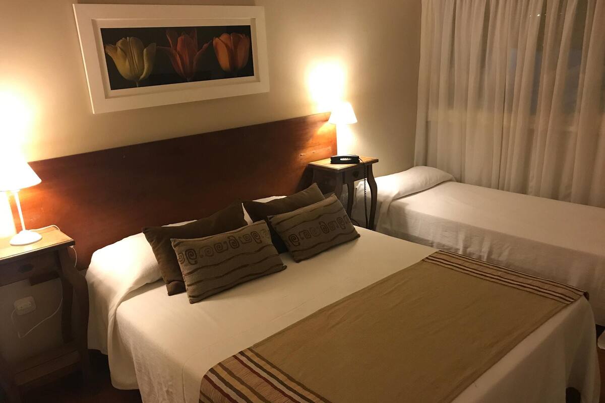 Quarto de hotel com duas camas, mesa de cabeceira com abajur, um quadro na parede e janela com cortinas.