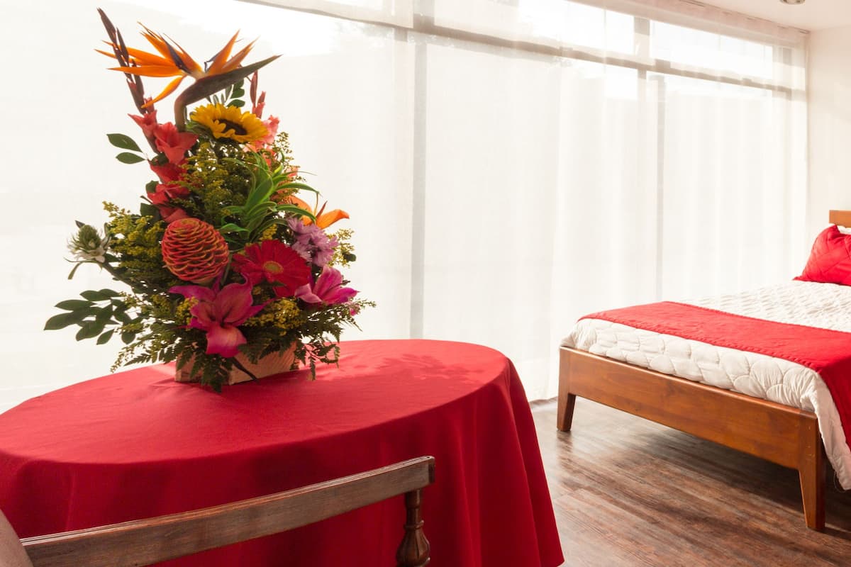 Une chambre d’hôtel avec deux lits, une tête de lit en bois, une table de chevet et une table recouverte d’une nappe rouge, ainsi qu’un arrangement floral.