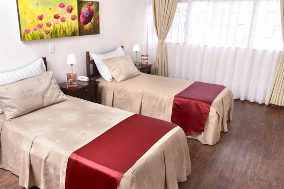 Une chambre d’hôtel avec deux lits, chacun recouvert d’une courtepointe beige et agrémenté d’un chemin de lit rouge, une table de chevet avec une lampe et un tableau représentant des fleurs accroché au mur.