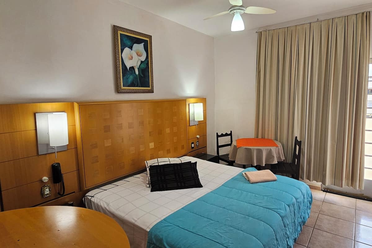 Une chambre d’hôtel avec un lit, une table, une chaise, un ventilateur de plafond, une fenêtre avec des rideaux et un tableau au mur.