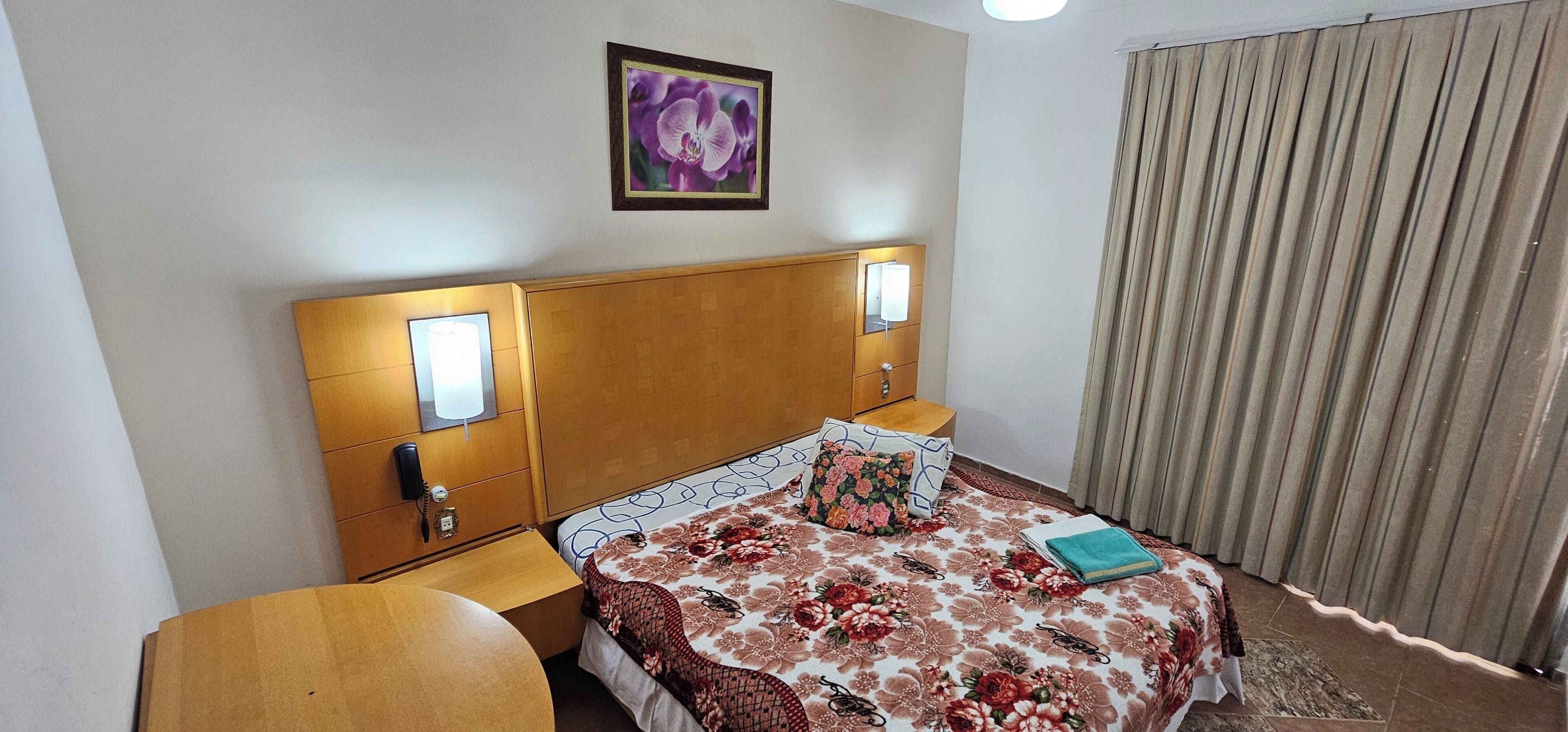 A hotel room with a wooden headboard, two bedside lamps, a floral bedspread, a nightstand, and a curtain.