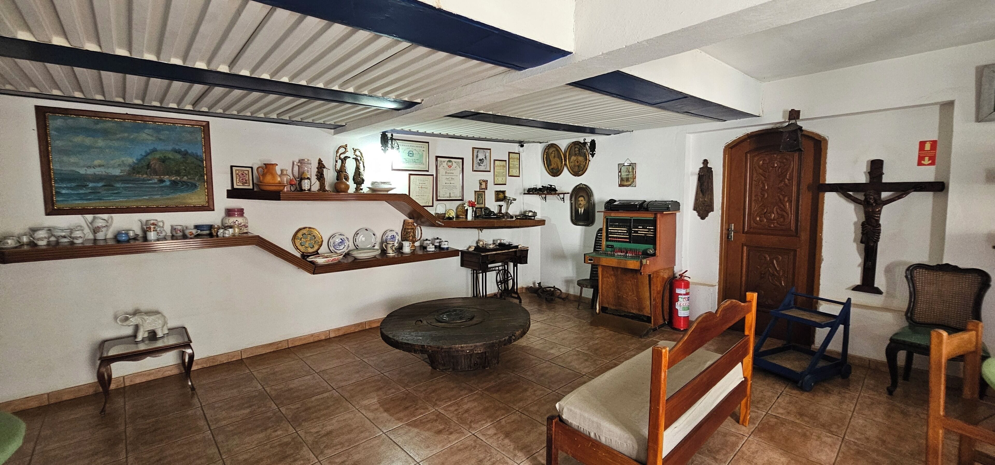 A room with a wooden bench, a small table, a shelf with various items, a painting, and a wooden door with a crucifix.
