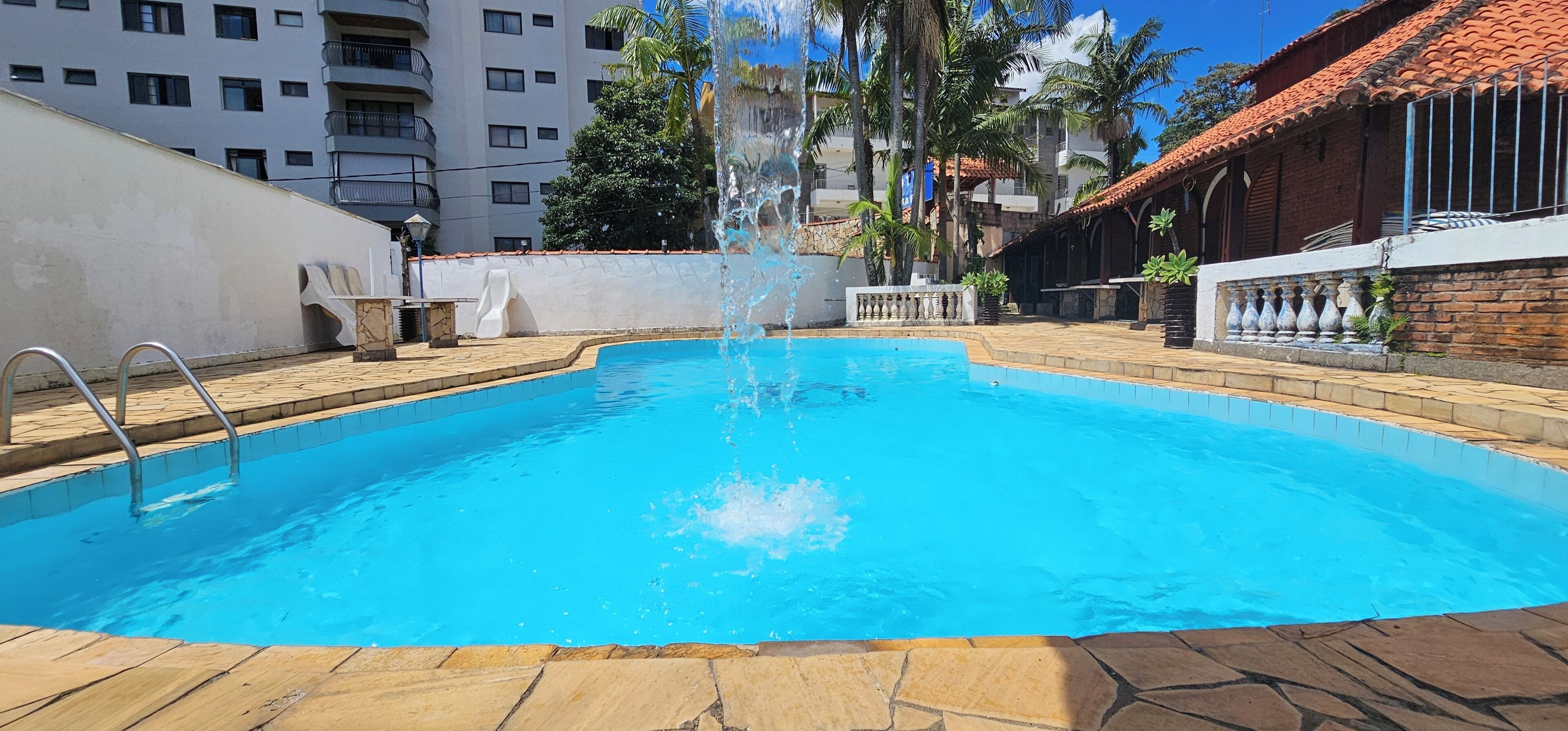A swimming pool with a water fountain, surrounded by a stone deck and a building with a tiled roof.
