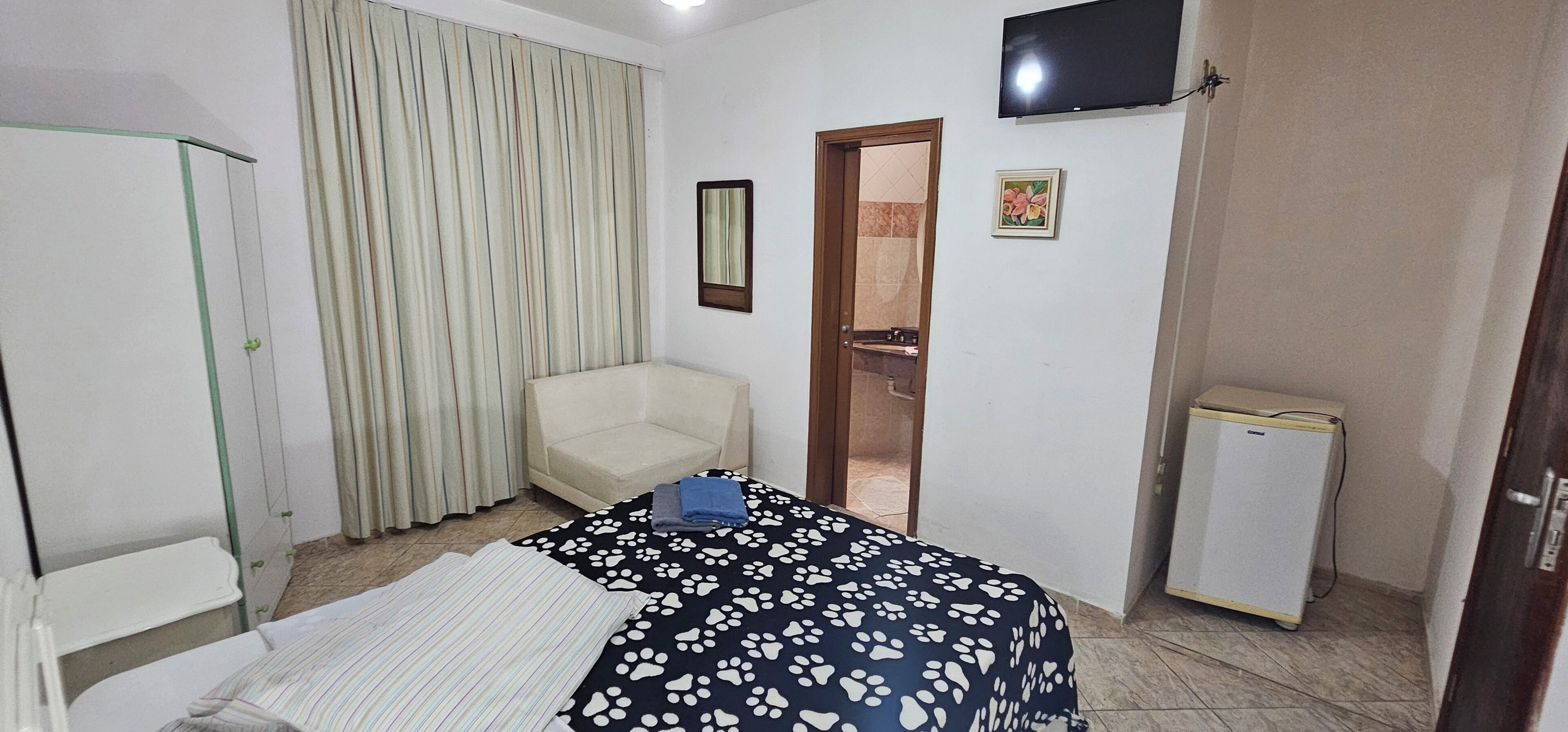 A hotel room with a bed, a chair, a television, a refrigerator, and a door leading to a bathroom.