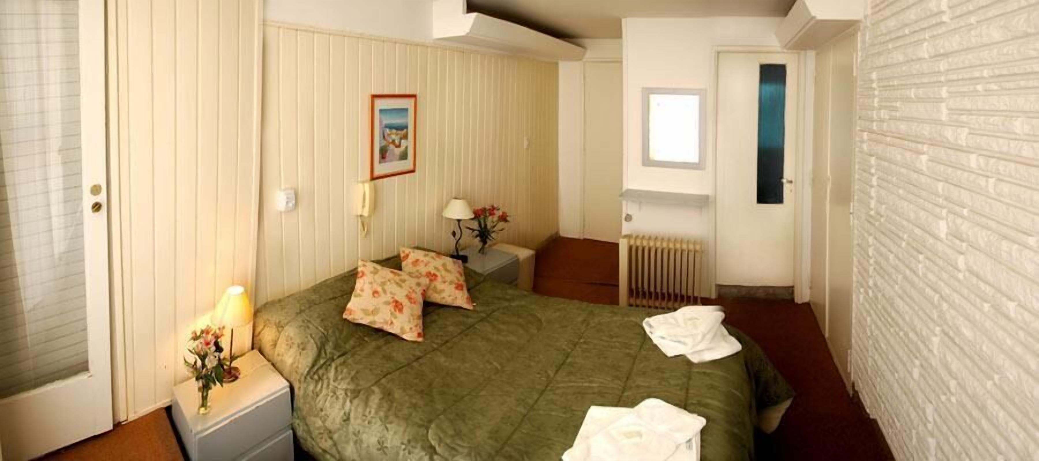 A bedroom with a green bedspread, a bedside table with a lamp, a framed picture on the wall, and a window with a view.